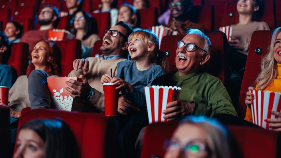 Spotlight on Father, daughter and grandfather laughing whilst watching a movie at the cinema eating popcorn. 